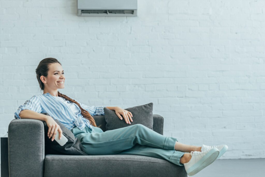 happy woman relaxing on sofa with air conditioner on white wall, summer heat concept